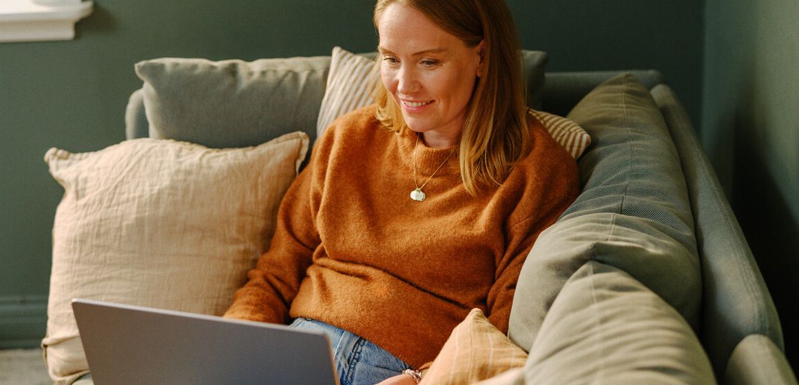 Kvinne i oransje genser sitter på sofaen med en laptop. Hun kan bruke Streamix for å spare penger.