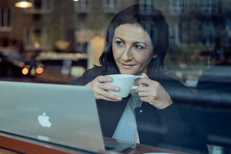 Dame ser ut av vindu mens hun drikker kaffe og jobber foran en PC